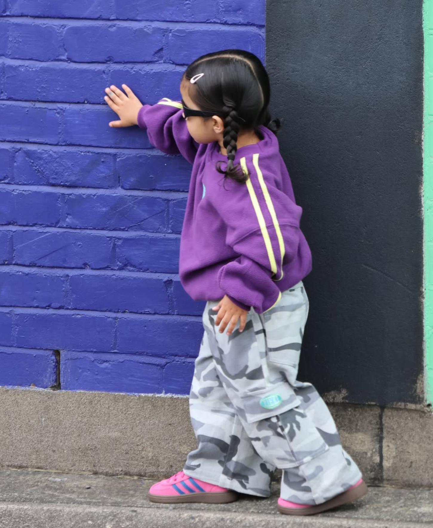 Toddler in purple sweatshirt and grey camo cargos by a purple brick wall